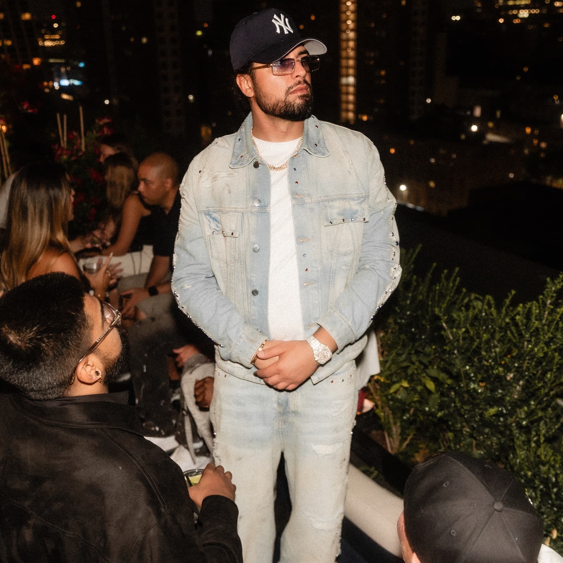 David Chaboki (Shibo / GodsBurnt) standing on a DDNYC 2026 rooftop in crystal denim and a Yankees cap in New York